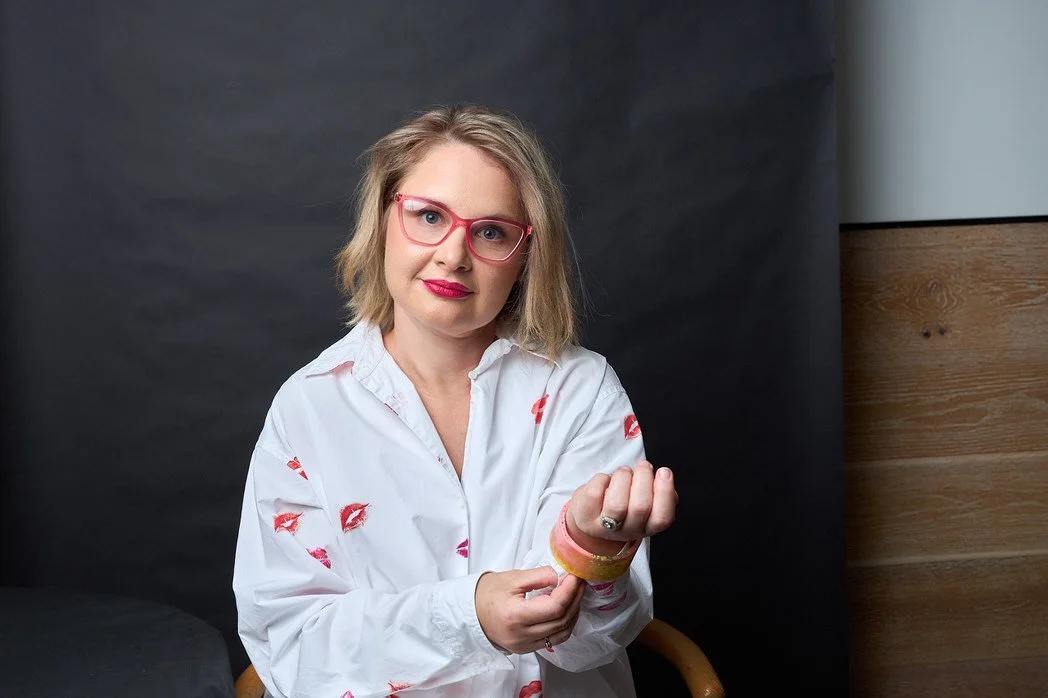 A woman with shoulder-length blonde hair, wearing pink glasses and a white shirt with red kiss marks, is sitting indoors against a dark background with a wooden panel on the right. She is adjusting her sleeve and looking at the camera.