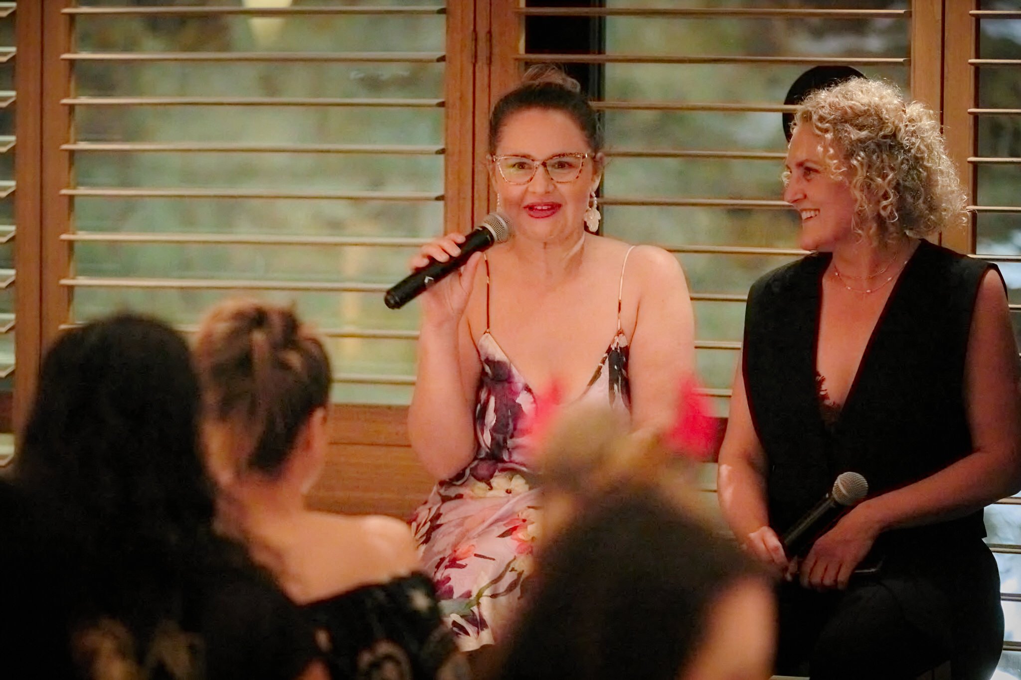 Two women sitting at a table during an event, with one woman speaking into a microphone and smiling, the other woman smiling and looking at her, with people in the foreground and a wooden wall with window shutters behind them.