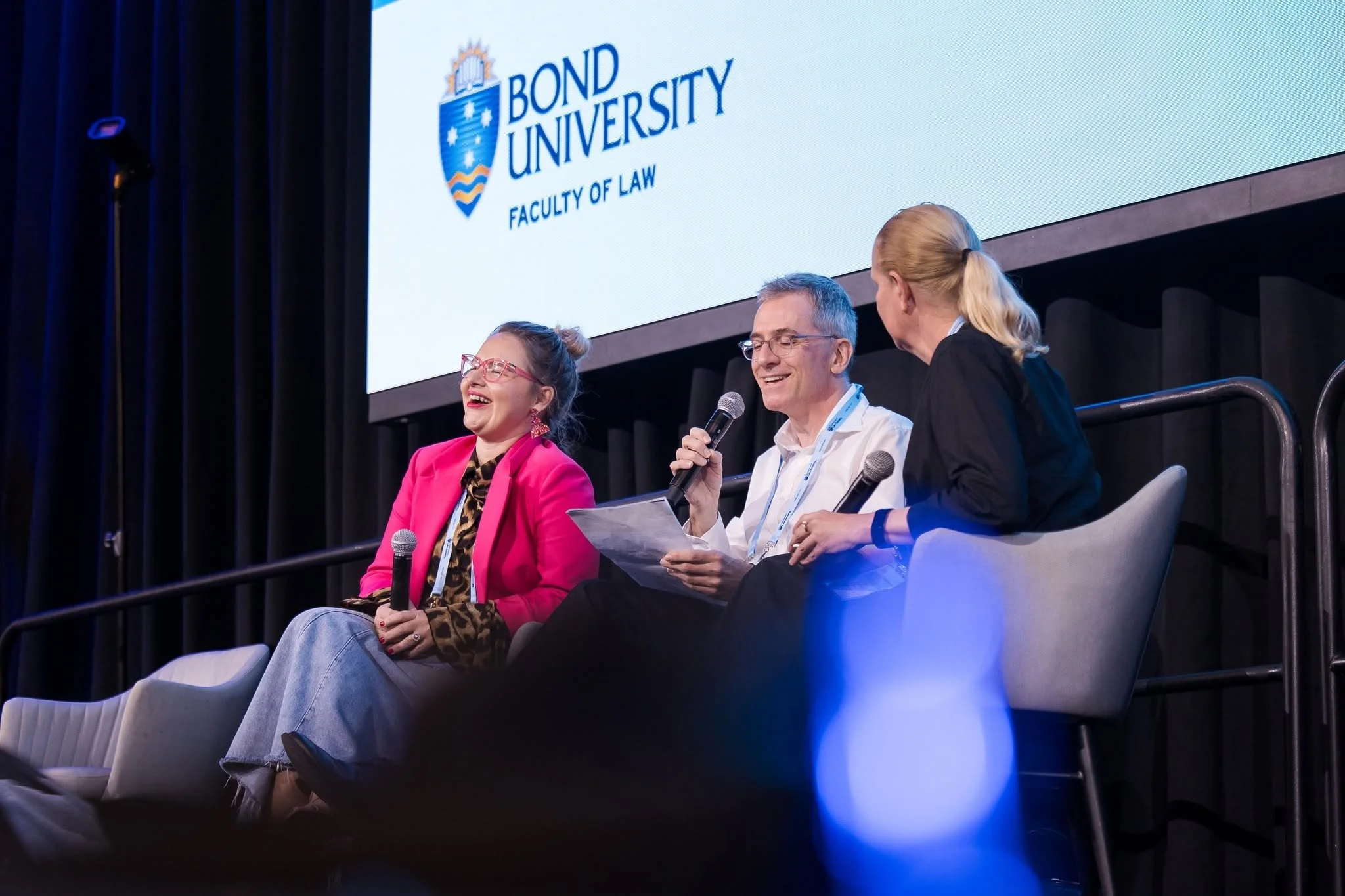 Three people sitting on a stage panel, speaking and listening with microphones, at a conference with a large screen displaying the "Bond University Faculty of Law" logo.  Leadership keynote speaker motivational speaker burnout recovery Rachael Karlyl