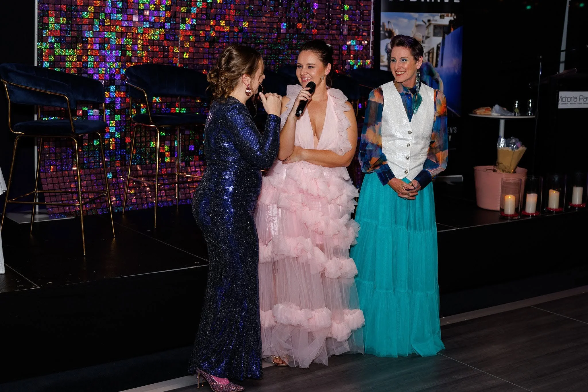 Three women are standing and talking, with one woman holding a microphone. The woman on the left has curly hair and is wearing a sparkly dark blue dress. The woman in the middle is wearing a pink ruffled gown, and the woman on the right has short hai