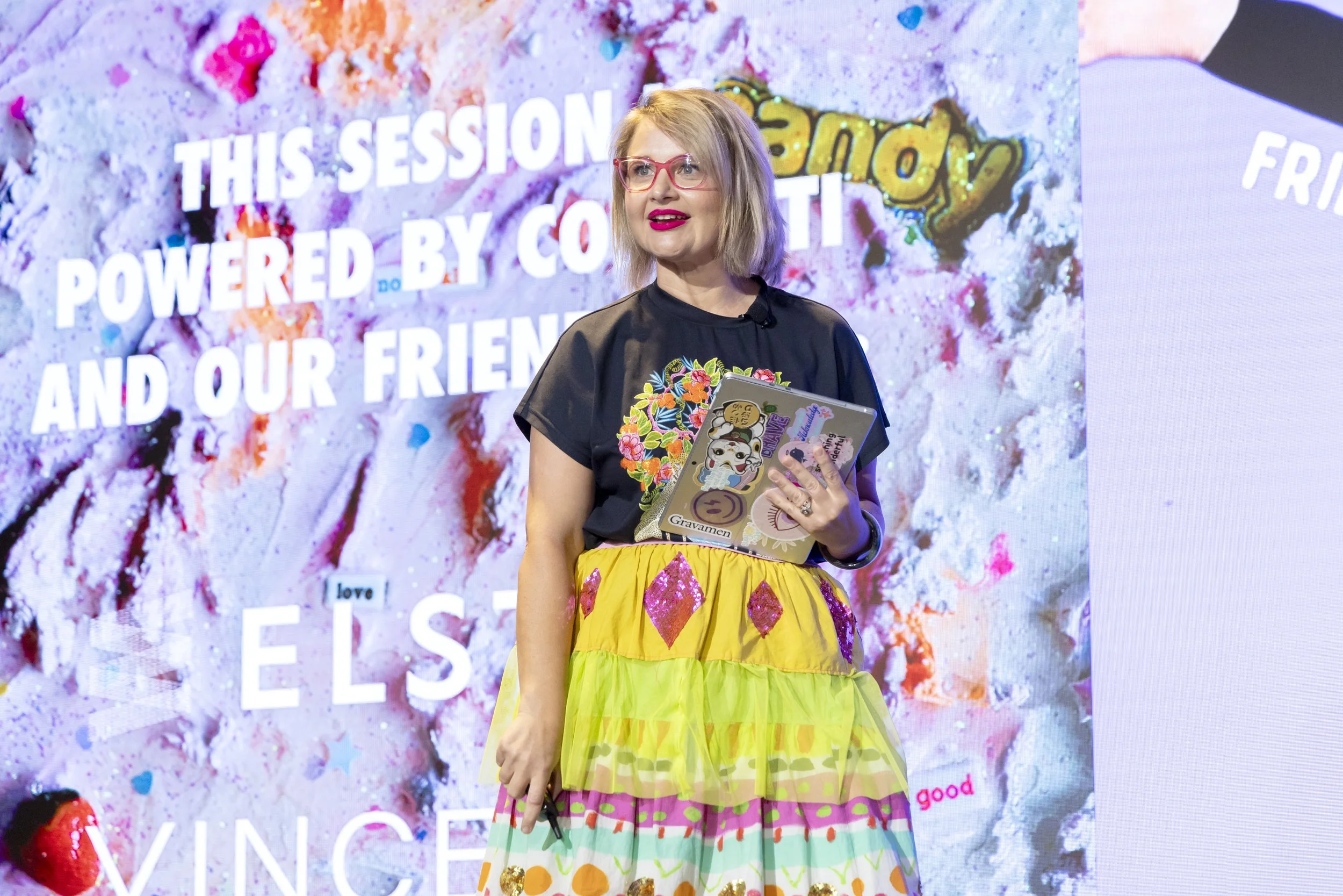 A woman with blonde hair wearing pink glasses and a black top with floral embroidery, holding a decorated laptop, stands in front of a colorful background with text that says 'This session powered by co...' and various colorful artwork.