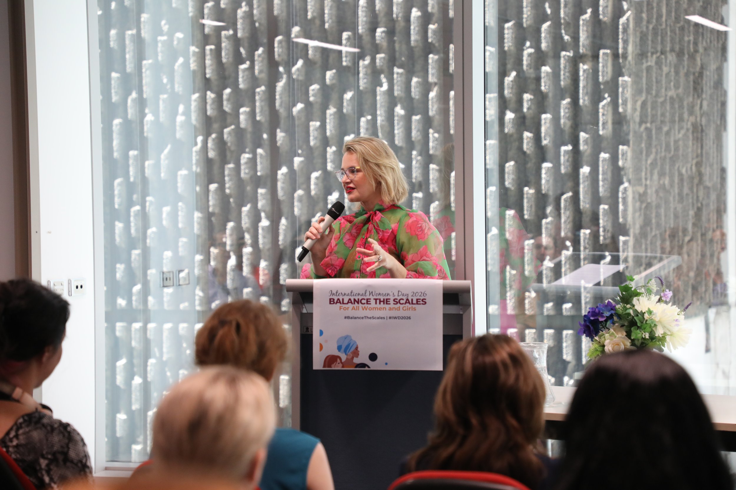 A woman with blonde hair and glasses speaking into a microphone at a podium during an International Women's Day event. The podium has a sign that reads 'Balance the Scales for All Women and Girls' with a colorful illustration. Audience members are se