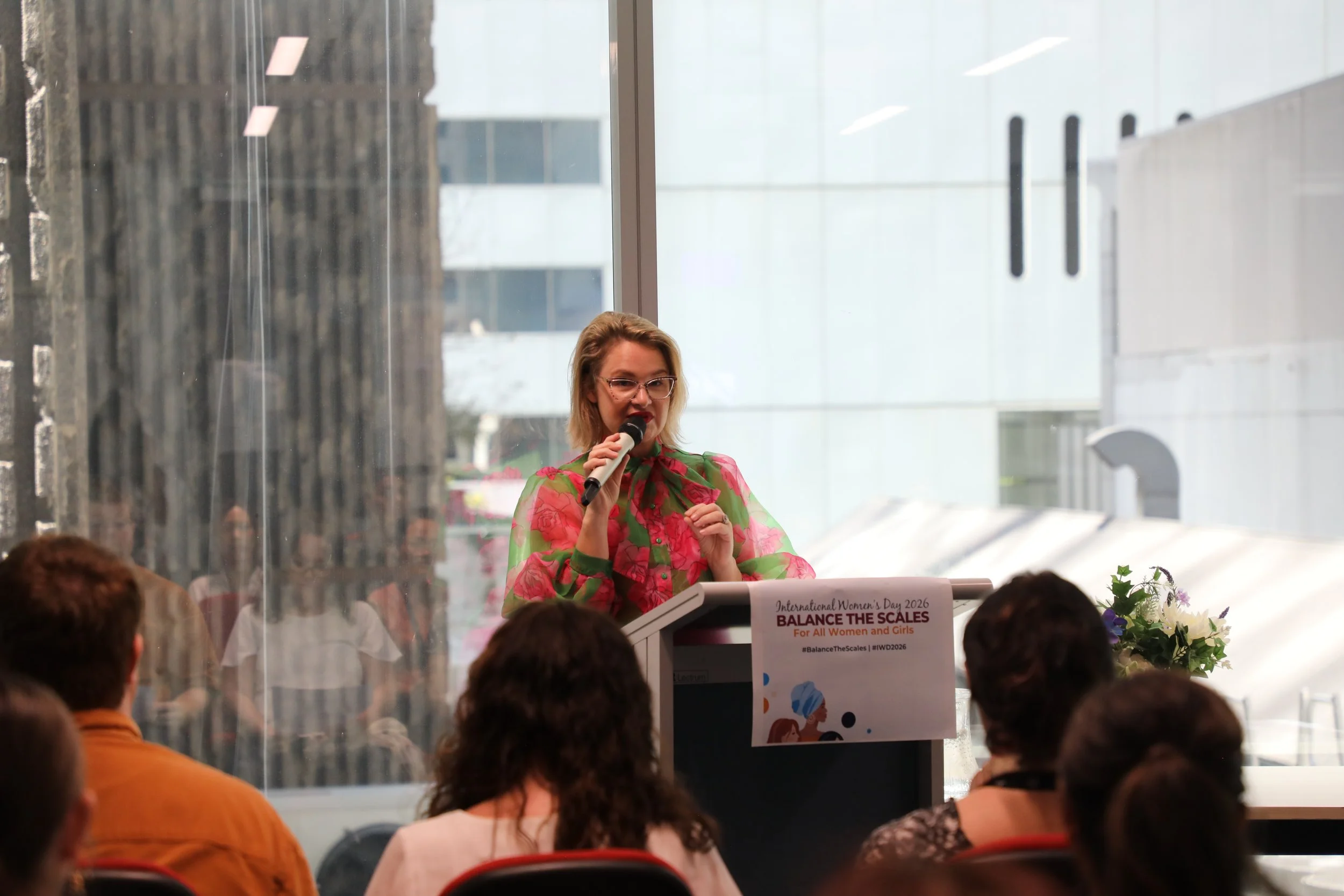 A woman with glasses and blonde hair speaking into a microphone at an indoor event, wearing a pink and green floral dress, with a sign promoting International Women's Day 2026, 'Balance the Scales for All Women and Girls', and an audience seated in f