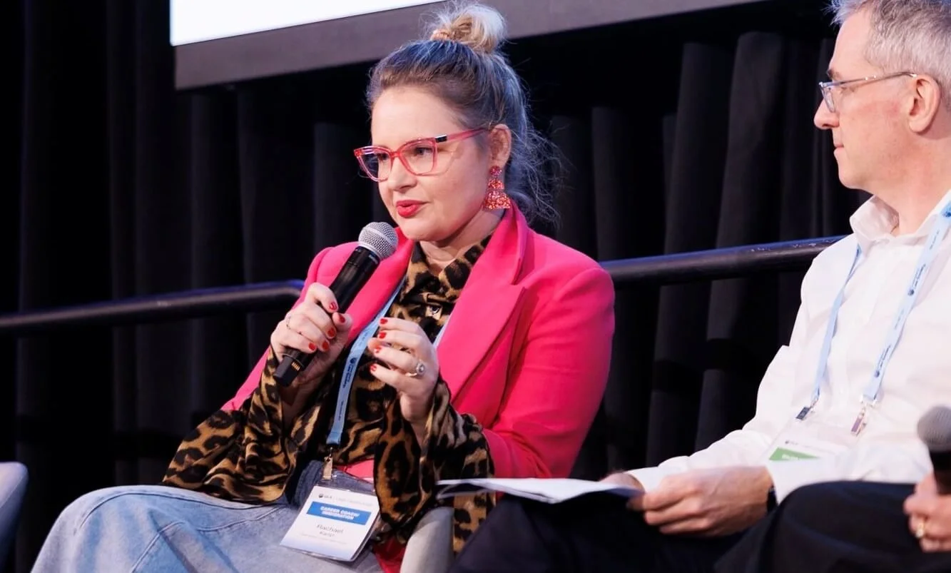 A woman wearing pink glasses, a pink blazer, and leopard print scarf speaking into a microphone at a panel discussion, with a man sitting beside her holding papers. Queensland law society  Leadership keynote speaker motivational speaker burnout recov