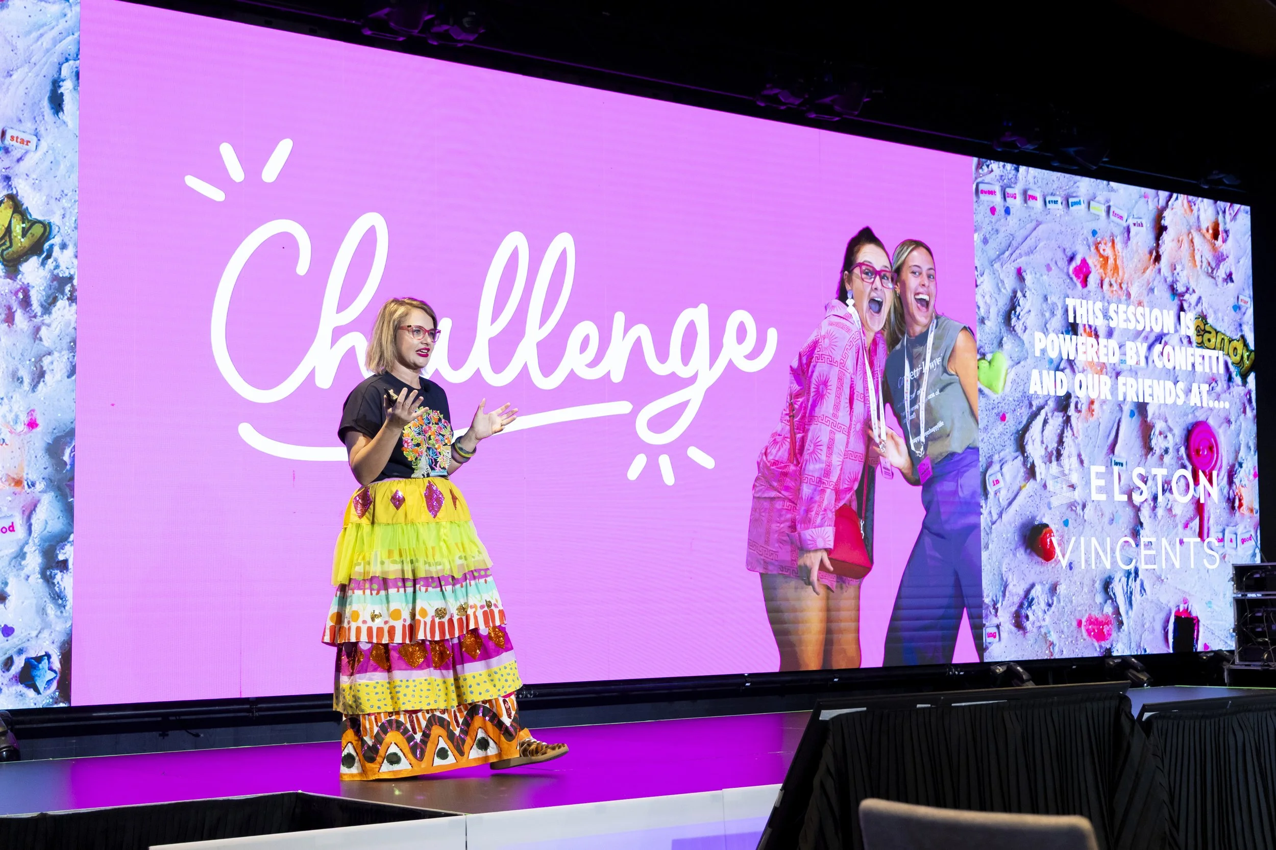 Woman dressed in a colorful, patterned skirt and black top standing on stage, speaking to an audience during a presentation or conference.