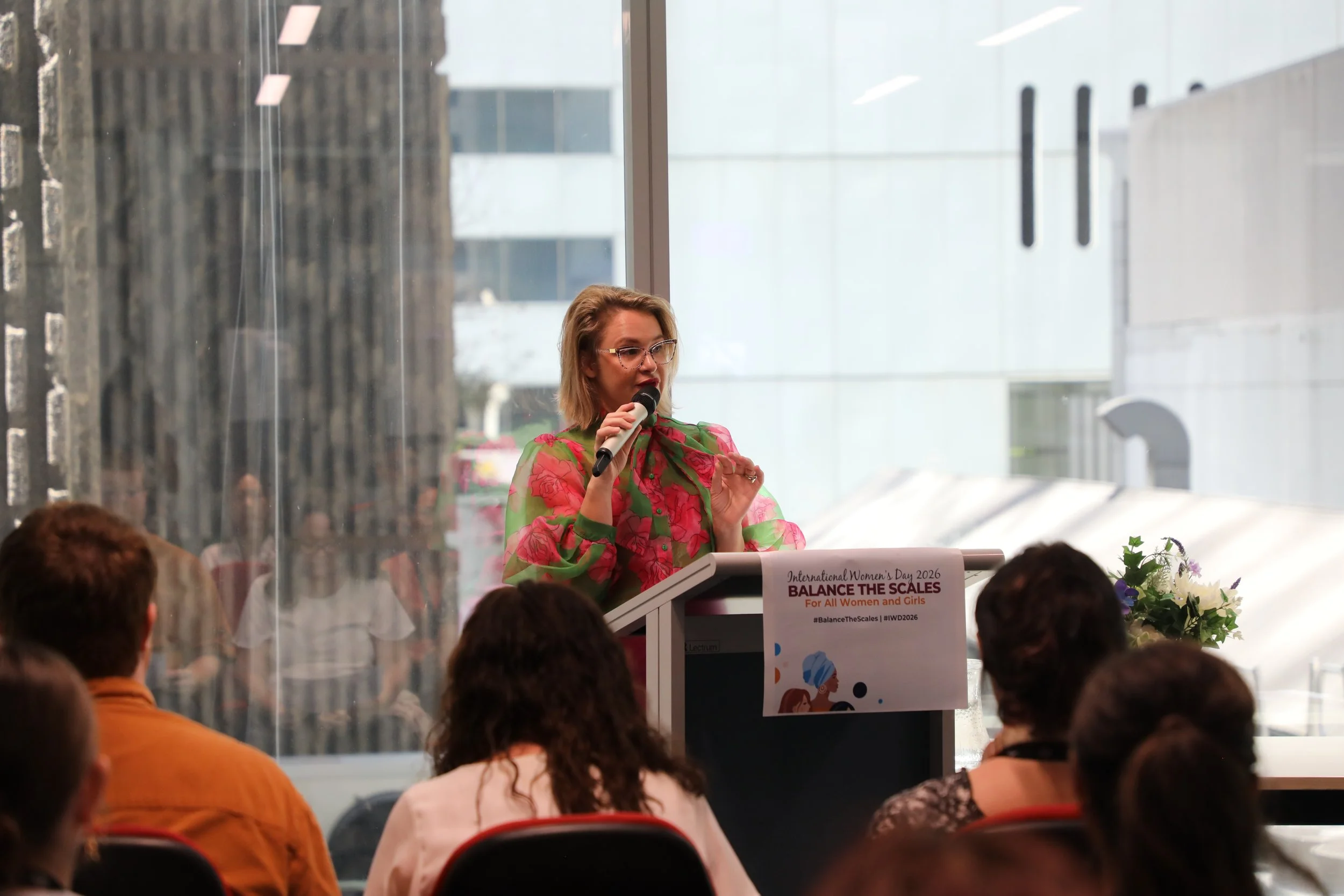 A woman with glasses and blonde hair speaking into a microphone at a podium with a sign that reads "International Women's Day 2026 Balance the Scales for All Women and Girls" during a daytime indoor event. Audience members are seated and visible in t