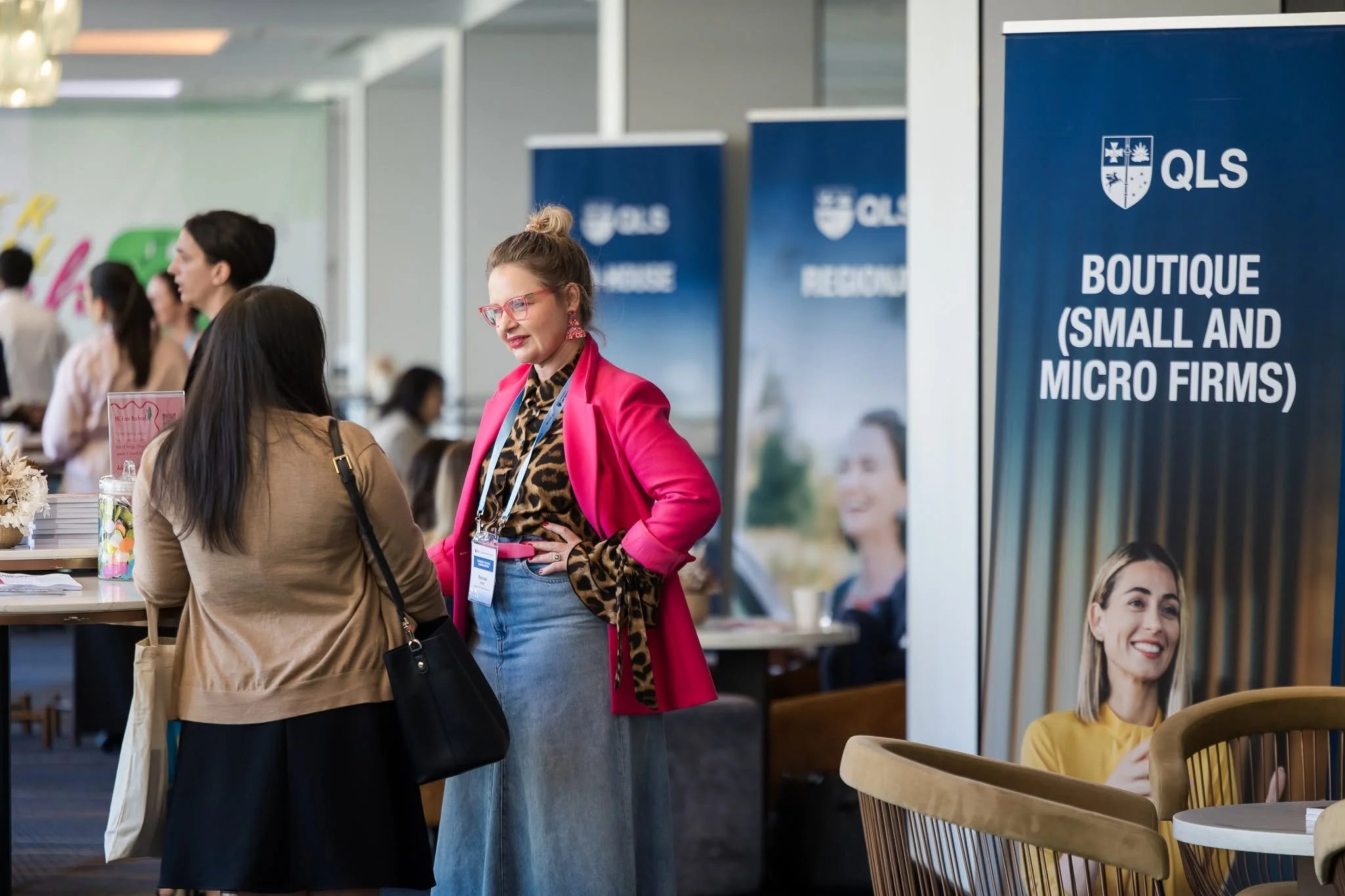 Two women are engaged in conversation at a conference or event, with a backdrop indicating a small and micro firm boutique, and other people in the background. Queensland law society rachael karlyl leadership key note speaker