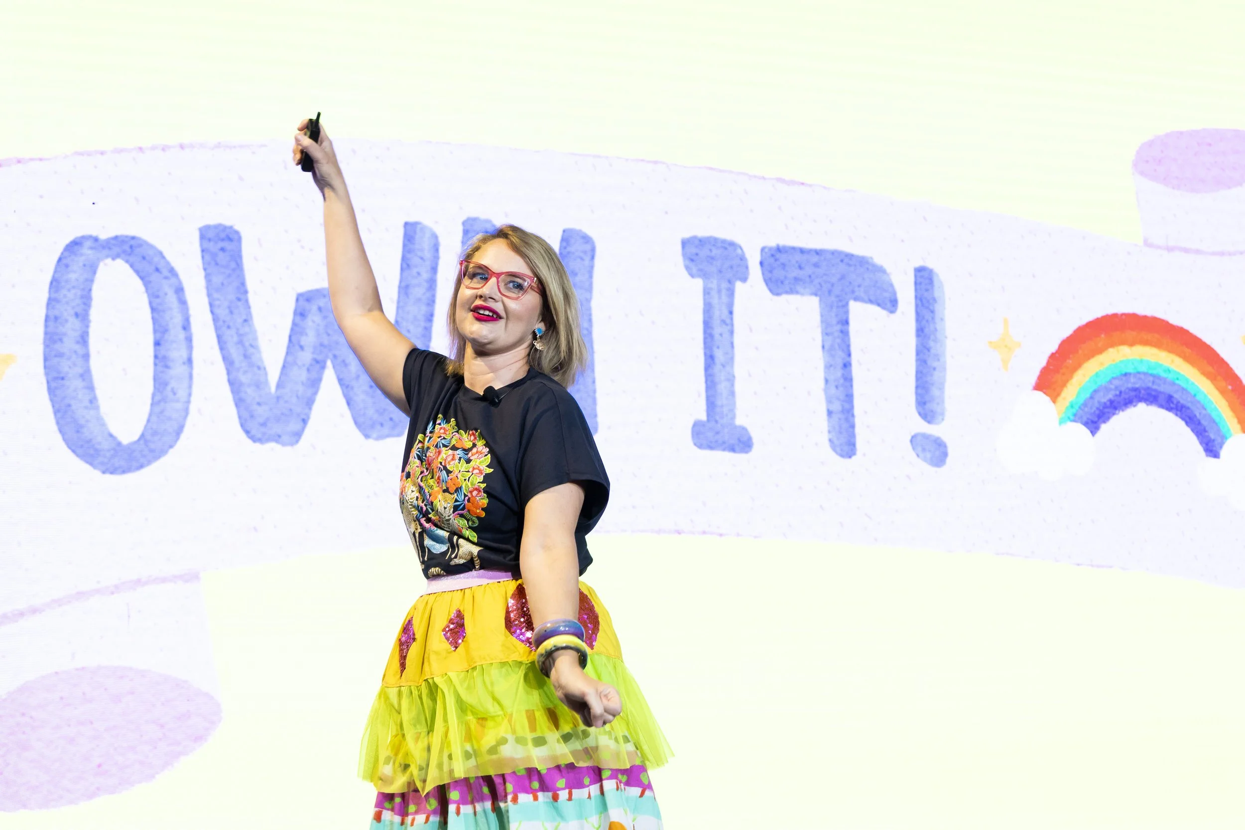 A woman with glasses and colorful earrings, wearing a black top with floral embroidery and a bright, multicolored, tiered skirt, is smiling and raising her right arm while standing on stage with a background display featuring a rainbow and the words 