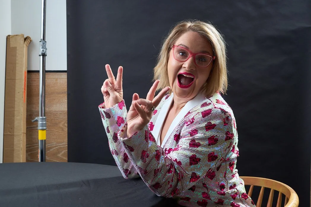 A woman with blonde hair, pink glasses, and red lipstick making a peace sign with both hands and smiling widely, sitting at a table with a black background.  Leadership keynote speaker motivational speaker burnout recovery Rachael Karlyl