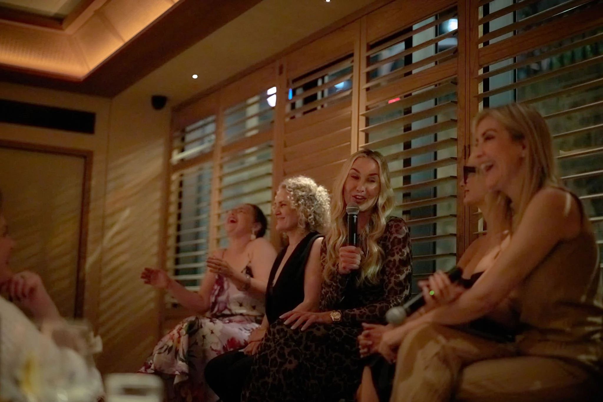 A group of women sitting in a wooden-paneled room, with one woman holding a microphone and speaking, while others are clapping and laughing. Connecting lawyer mums  Leadership keynote speaker motivational speaker burnout recovery Rachael Karlyl