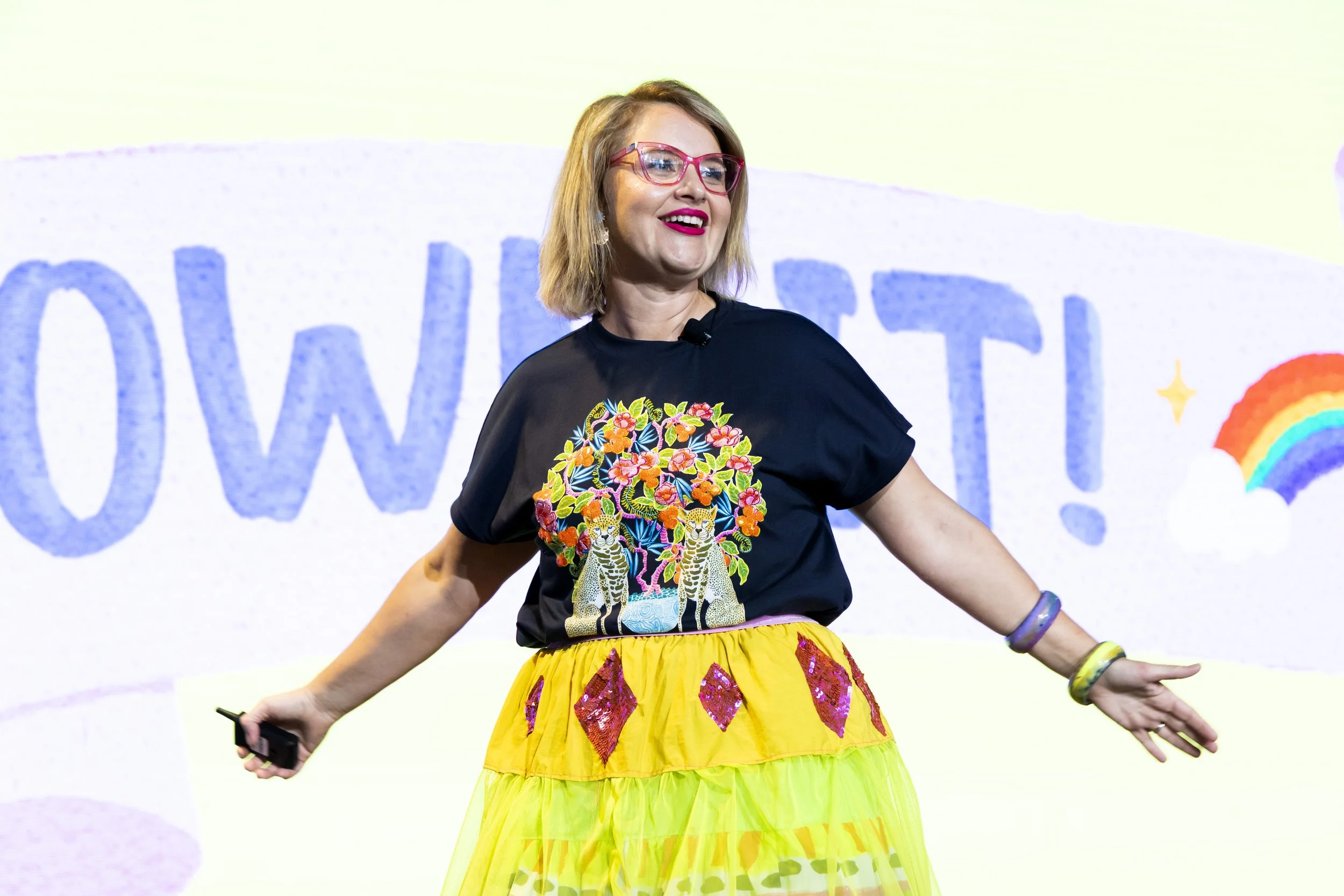 Woman wearing pink glasses and colorful black t-shirt with a tiger and floral print, smiling on stage, holding a microphone, yellow layered skirt with pink and purple accents, and rainbow and star in the background.