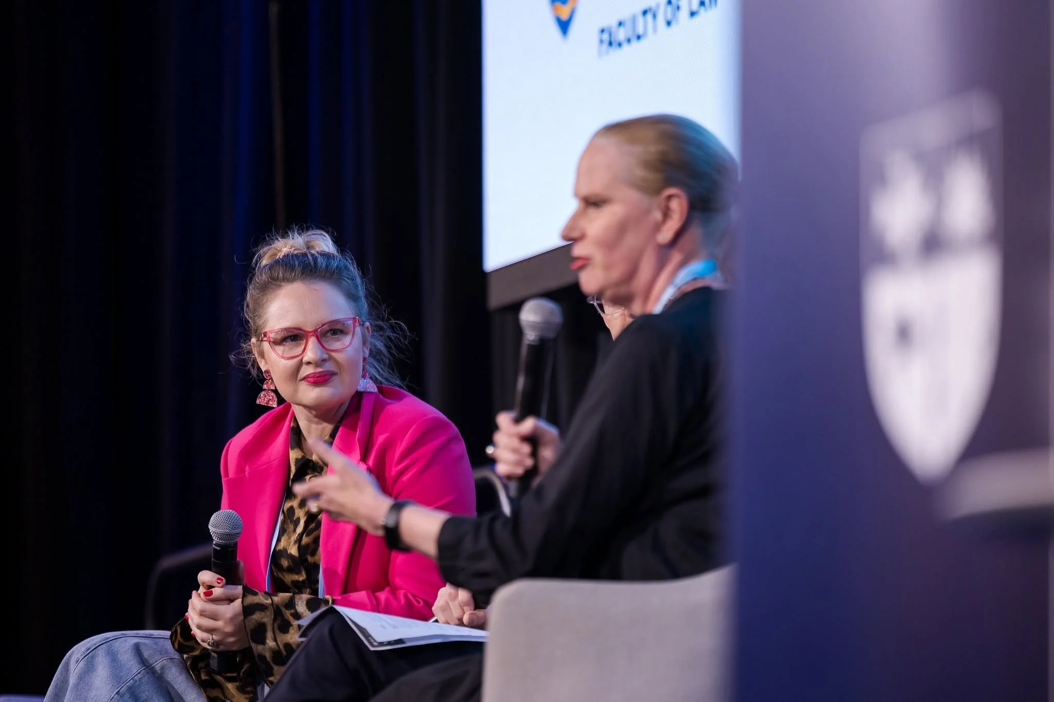 Two women sitting on stage engaged in conversation, both holding microphones. The woman on the left is wearing bright pink glasses, a pink blazer, and a leopard print shirt, listening attentively. The woman on the right has blonde hair in a bun, wear