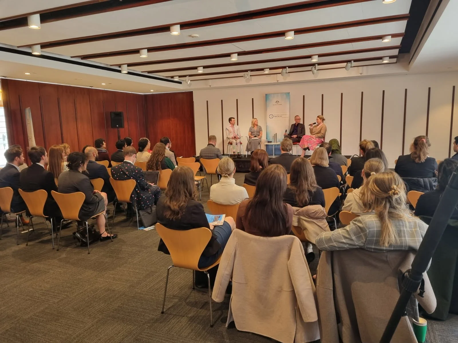A panel discussion with four people on a stage in front of an audience in a conference room. The audience is seated and listening attentively. Government lawyers. Australian Government legal service.  Leadership keynote speaker motivational speaker 
