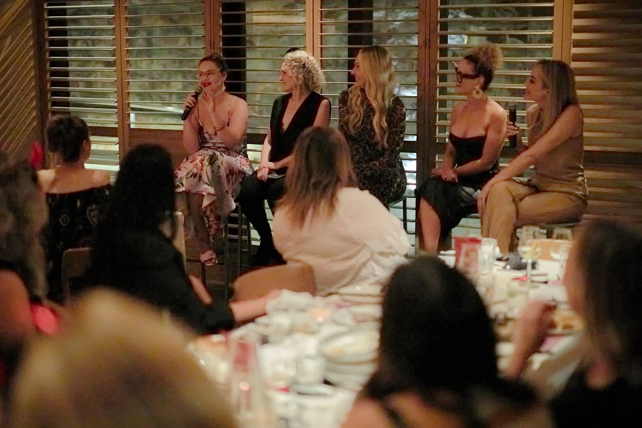 A group of five women sitting on a stage area in front of an audience, with wooden paneling and window shutters in the background. One woman is speaking into a microphone, while the others listen and smile.