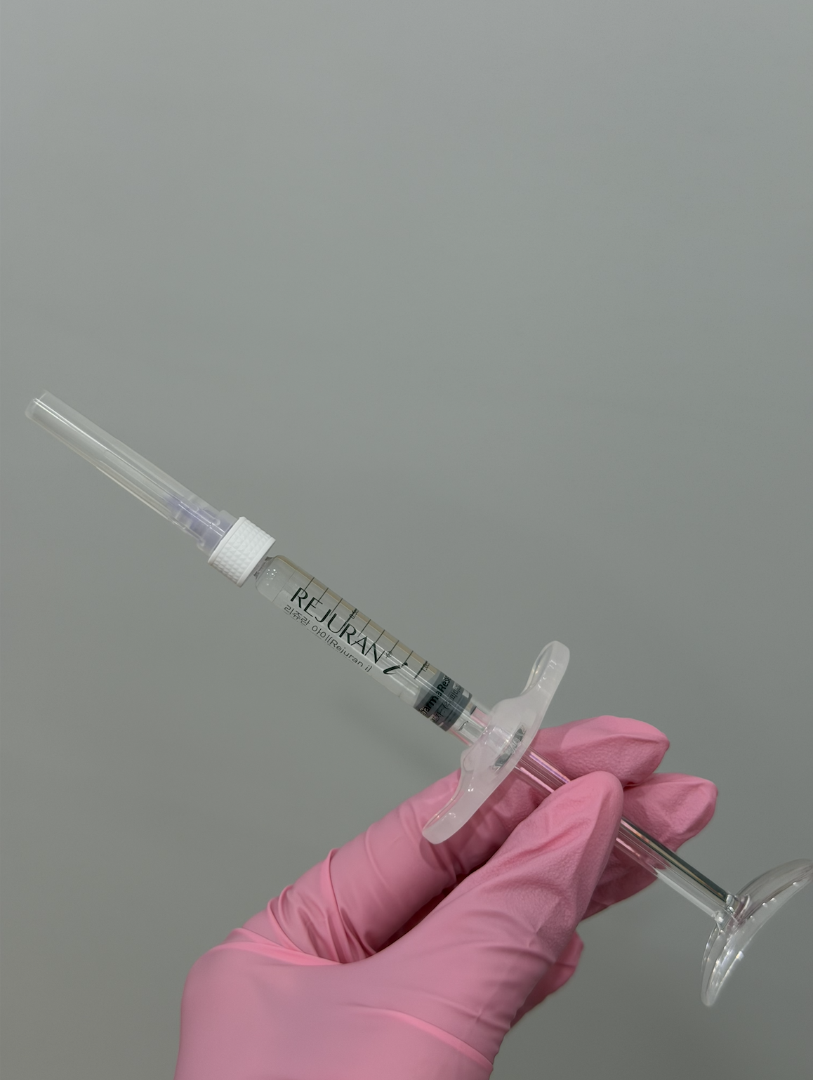 A hand wearing a pink glove holding a syringe with a clear liquid. The syringe has the label 'REJURAN' and appears to contain a cosmetic or medical solution. The background is plain and gray.