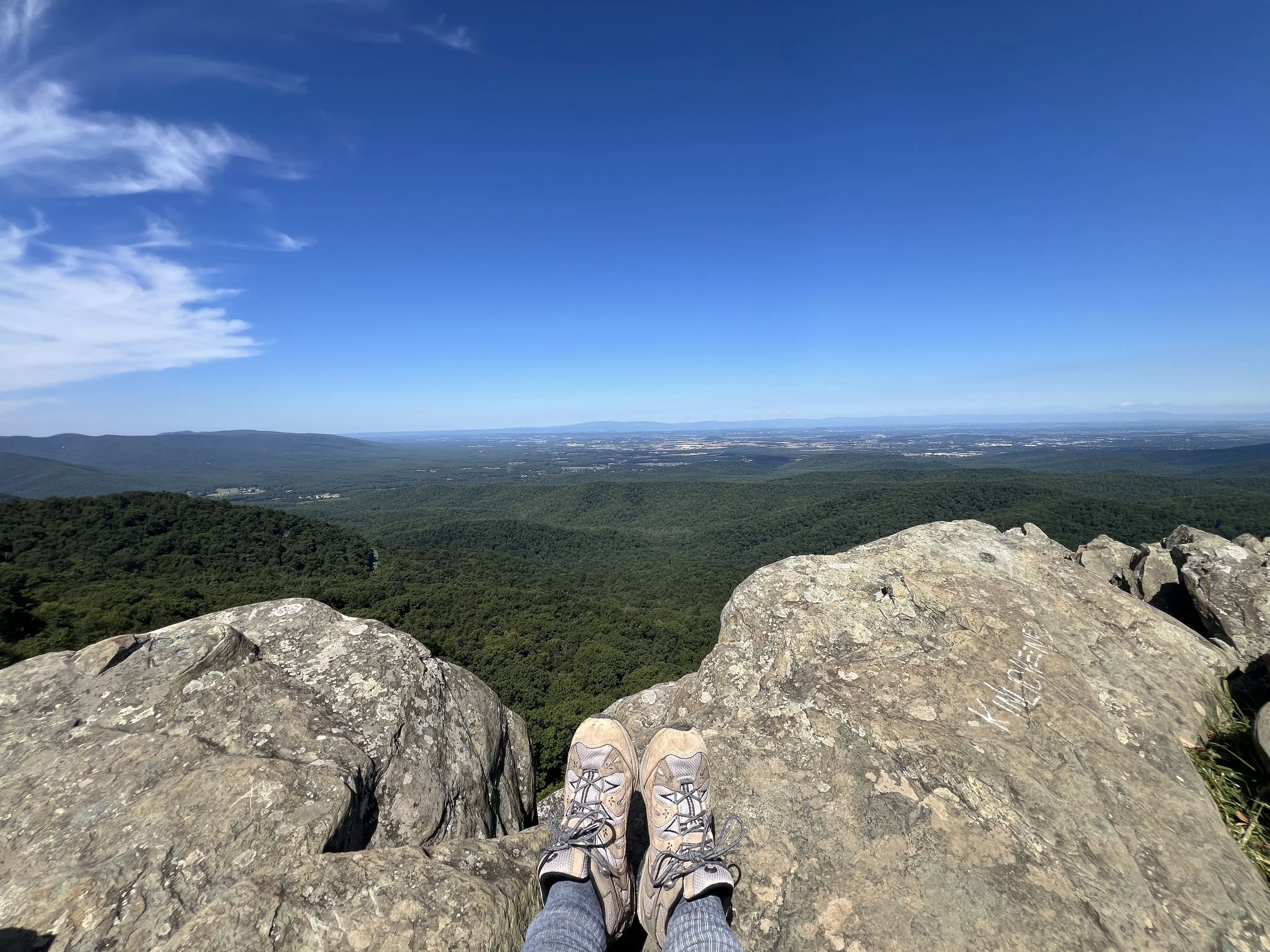 View from a mountain ledge with a person’s feet in hiking shoes, overlooking a vast landscape of forested hills, plains, and a blue sky with wispy clouds.