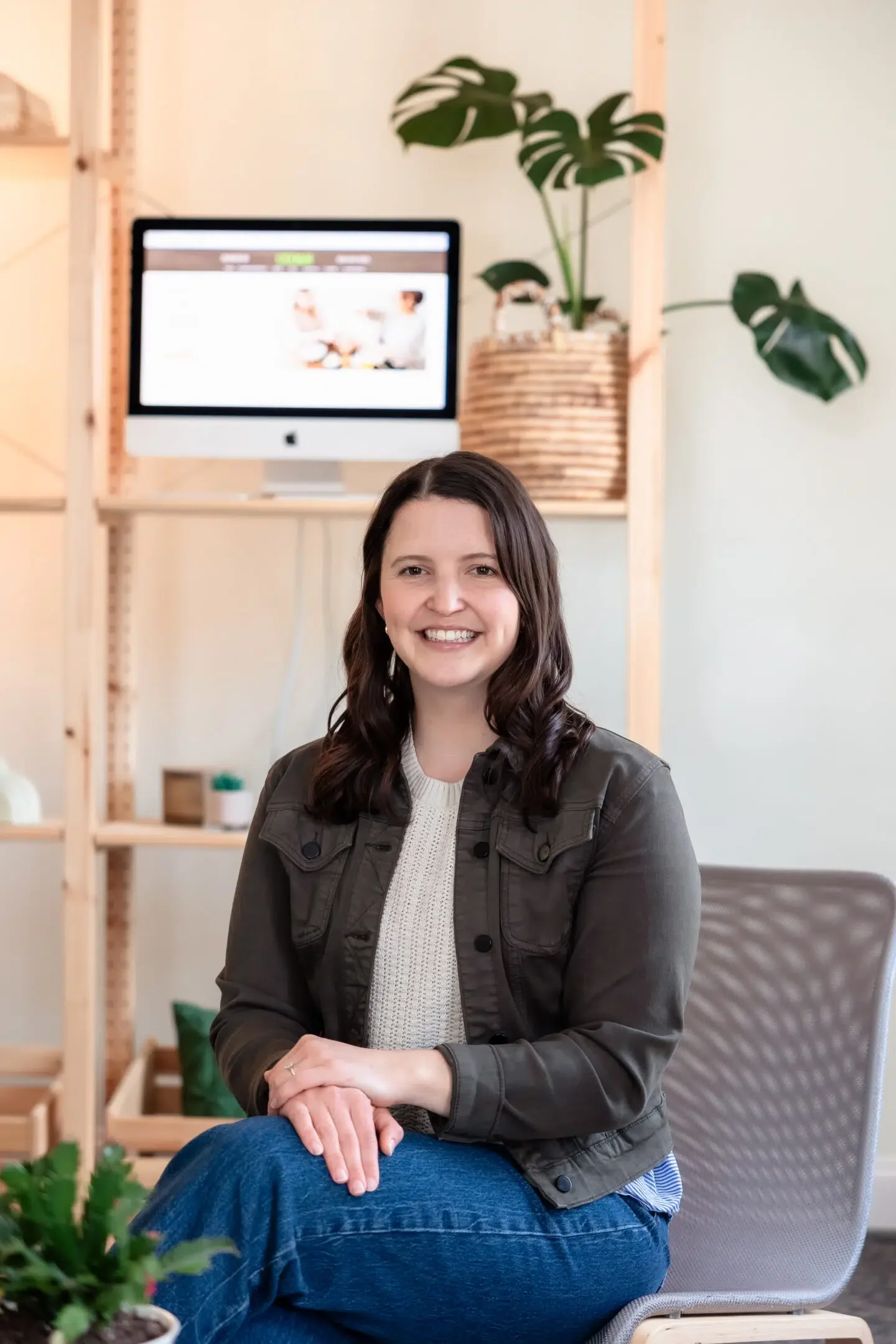 A smiling woman sitting in a modern office space with a computer monitor, plants, and wooden shelves in the background.