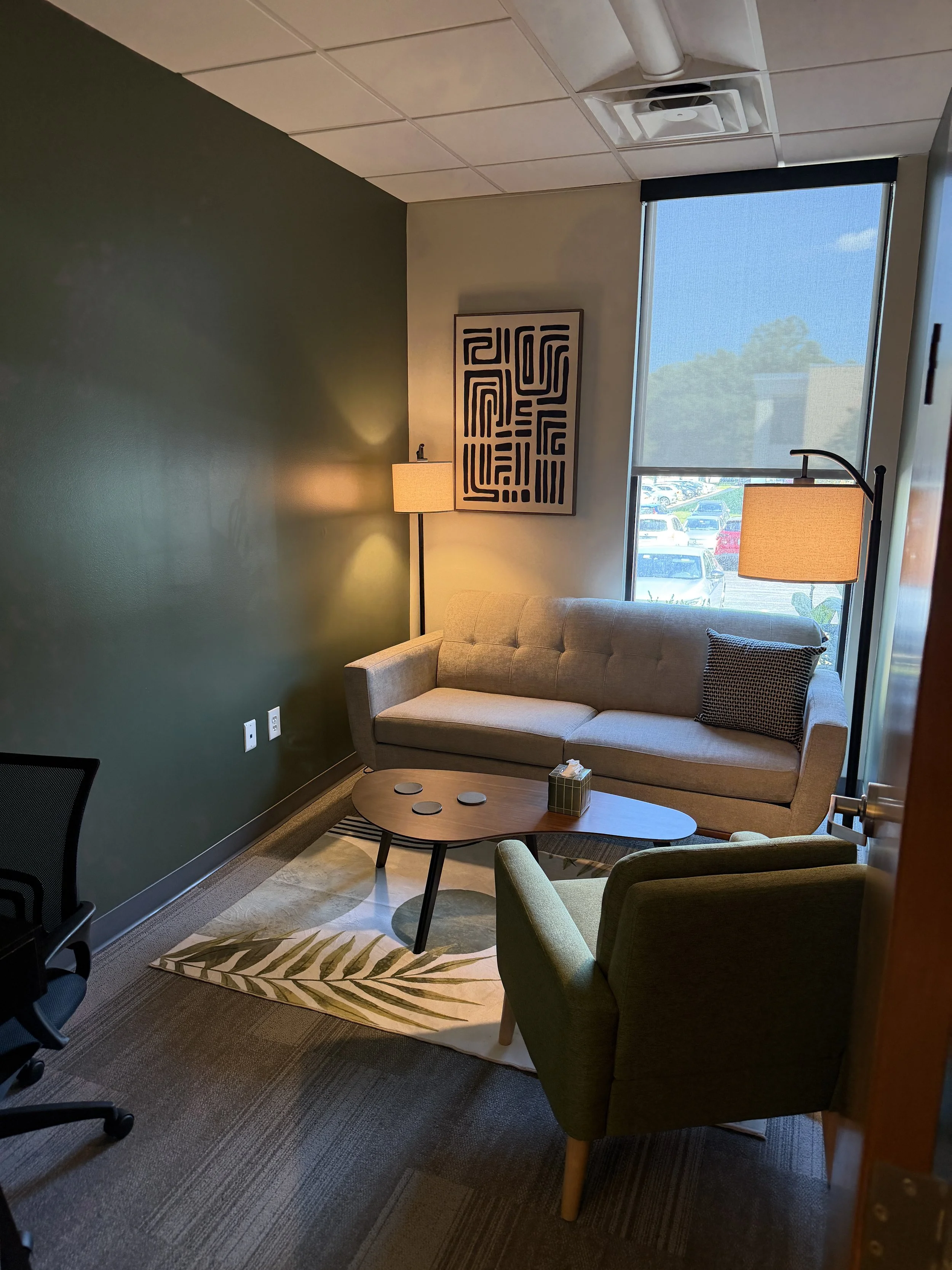 Office lounge with a beige sofa, green armchair, two floor lamps with beige shades, a round coffee table with coasters and a tissue box, a framed piece of abstract black and white art, a large window with a view of a parking lot and blue sky.