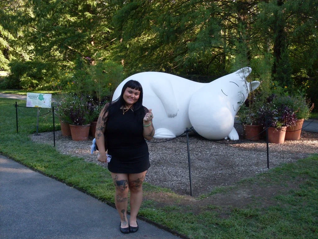 A woman with black hair, tattoos on her arms and legs, wearing a black dress, standing next to Moomin, surrounded by potted plants at the New York Botanical Gardens.