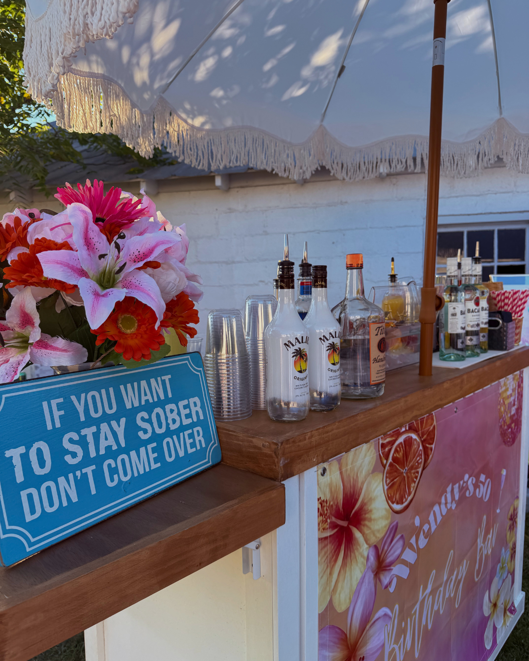 A wooden outdoor bar with bottles of alcohol, plastic cups, and floral decorations, including a pink and white flower arrangement. A blue sign on the stand reads, "If you want to stay sober don't come over." A large white umbrella provides shade.