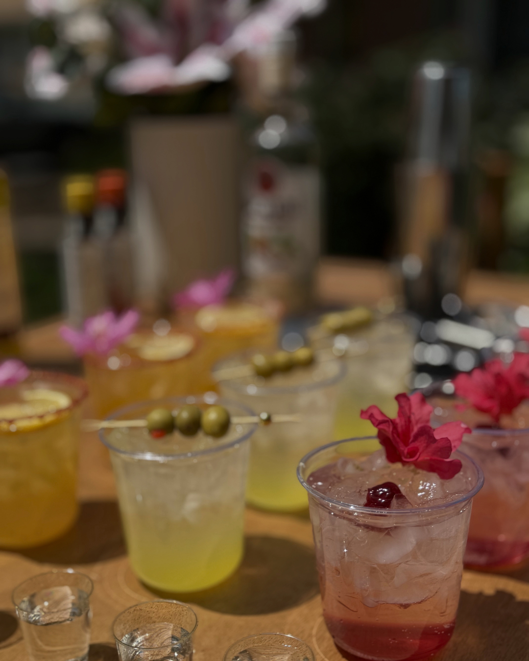 Assorted colorful cocktails with flowers and garnishes on a wooden surface, with blurred bottles in the background.