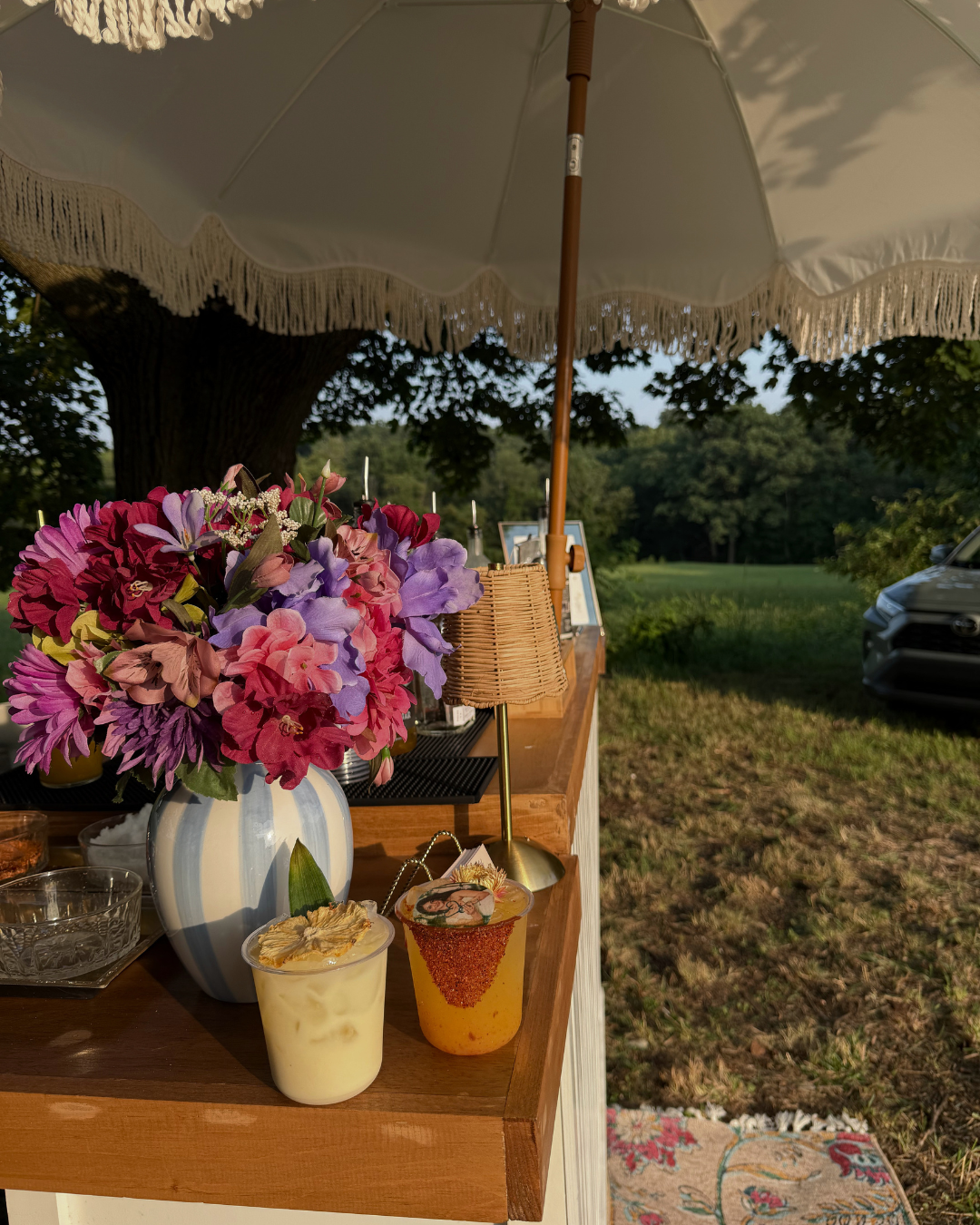 A floral arrangement with pink and purple flowers in a white vase, two drinks with garnishes, and a small wicker lamp on a wooden outdoor bar under a large white umbrella in a grassy outdoor setting.