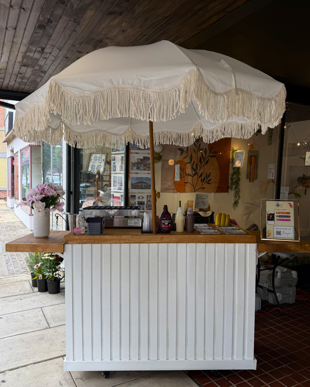 A small outdoor concession stand with a white fringed umbrella, a flower vase, and various condiment bottles, located beside a storefront with windows and interior artwork.