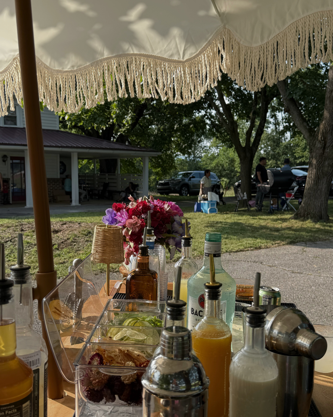 A shaded outdoor drinks table with booze, juices, lime slices, and a flower arrangement, with people socializing and cars in the background.