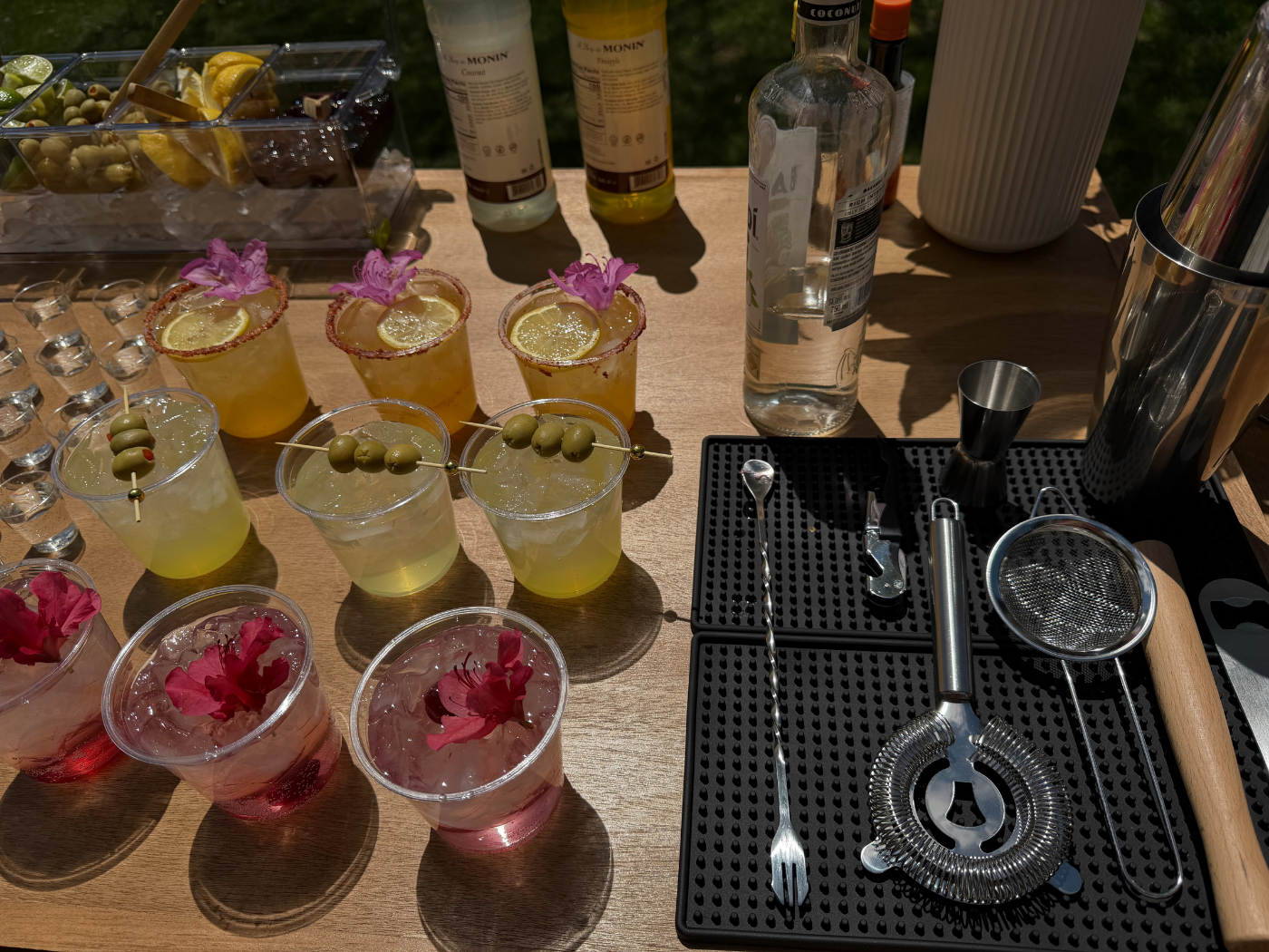An arrangement of colorful cocktails on a wooden table, topped with flowers, olives, and lemon slices, with bartending tools and bottles in the background.