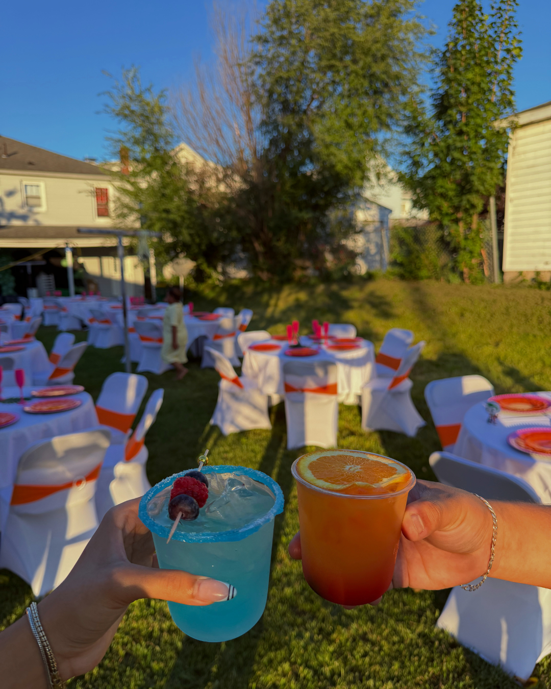 Two hands holding colorful cocktails in the foreground, outdoor party setup with round tables, white chairs, and decorations in the background on a sunny day.