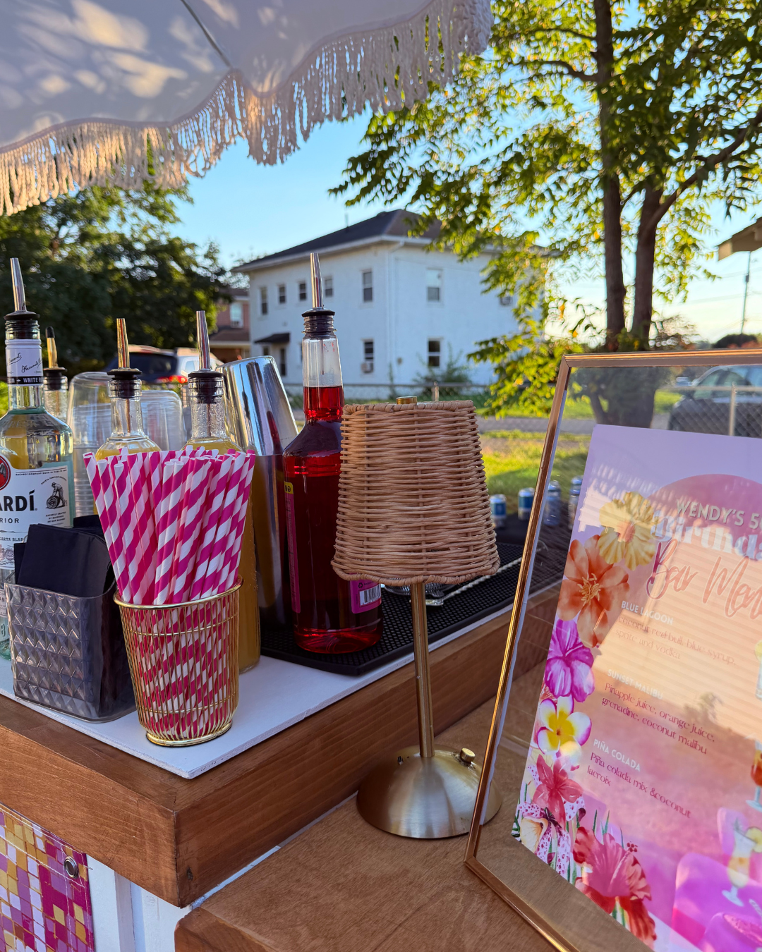 Bar setup with various bottles and striped straws, a sign with colorful floral design, an umbrella overhead, and a residential house in the background during sunset.