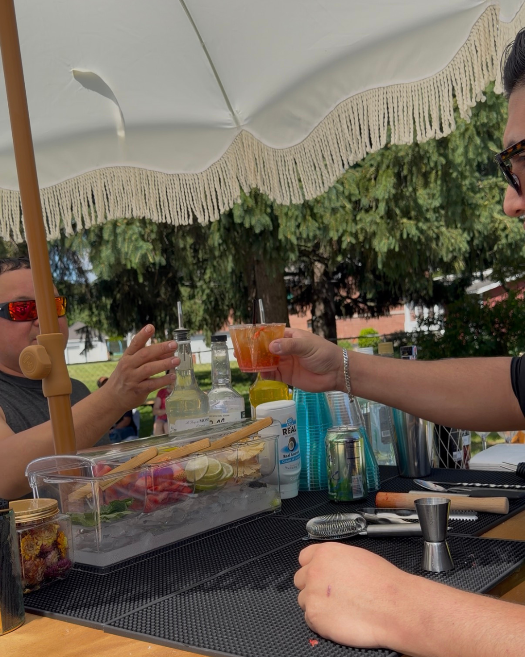A person with sunglasses is getting a cocktail handed by a bartender at an outdoor bar under a white umbrella on a sunny day.