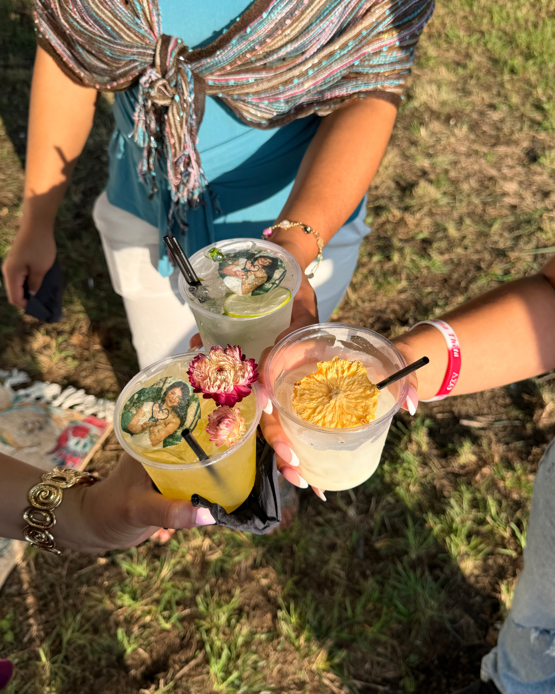 Three people holding plastic cups with drinks, garnished with pineapple, flowers, and edible custom toppers, outdoors on grassy ground.