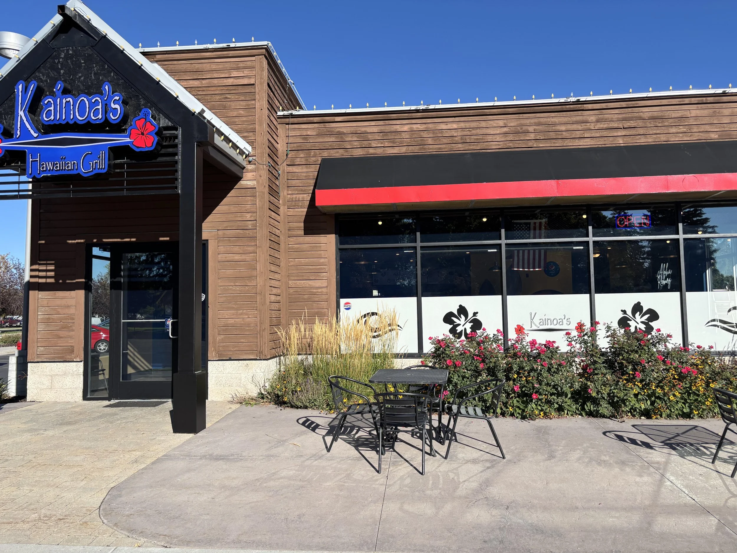 Exterior of Kaino's Hawaiian Grill restaurant with wood siding, large window with an 'Open' sign, plants and flowers outside, and outdoor seating area with metal tables and chairs.
