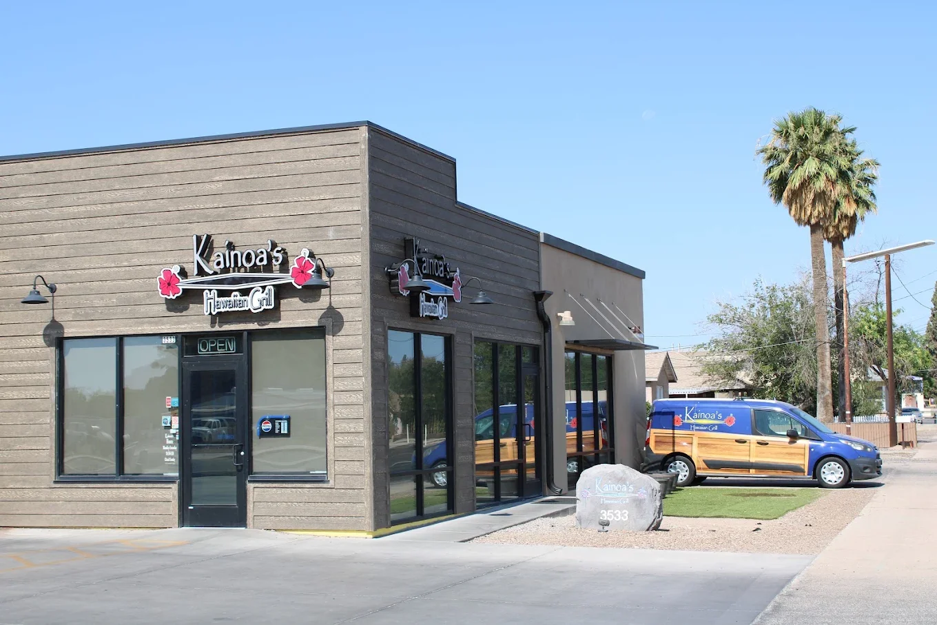 Exterior view of Kaino's Hawaiian Grill restaurant with a brown wood facade, large glass windows, and a Hawaiian-themed van parked outside, including palm trees and a clear blue sky.
