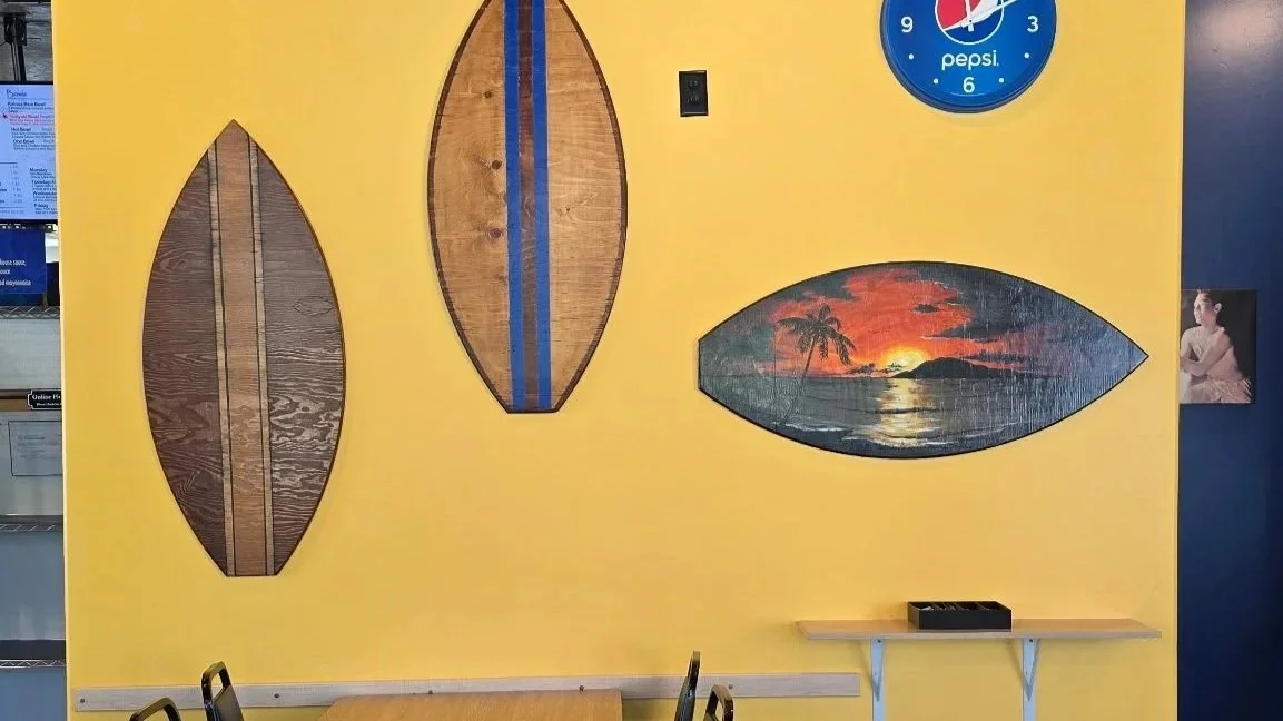 Decorative wooden surfboards and a tropical sunset painting on a yellow wall in a room with a blue wall, clock, and small shelf.