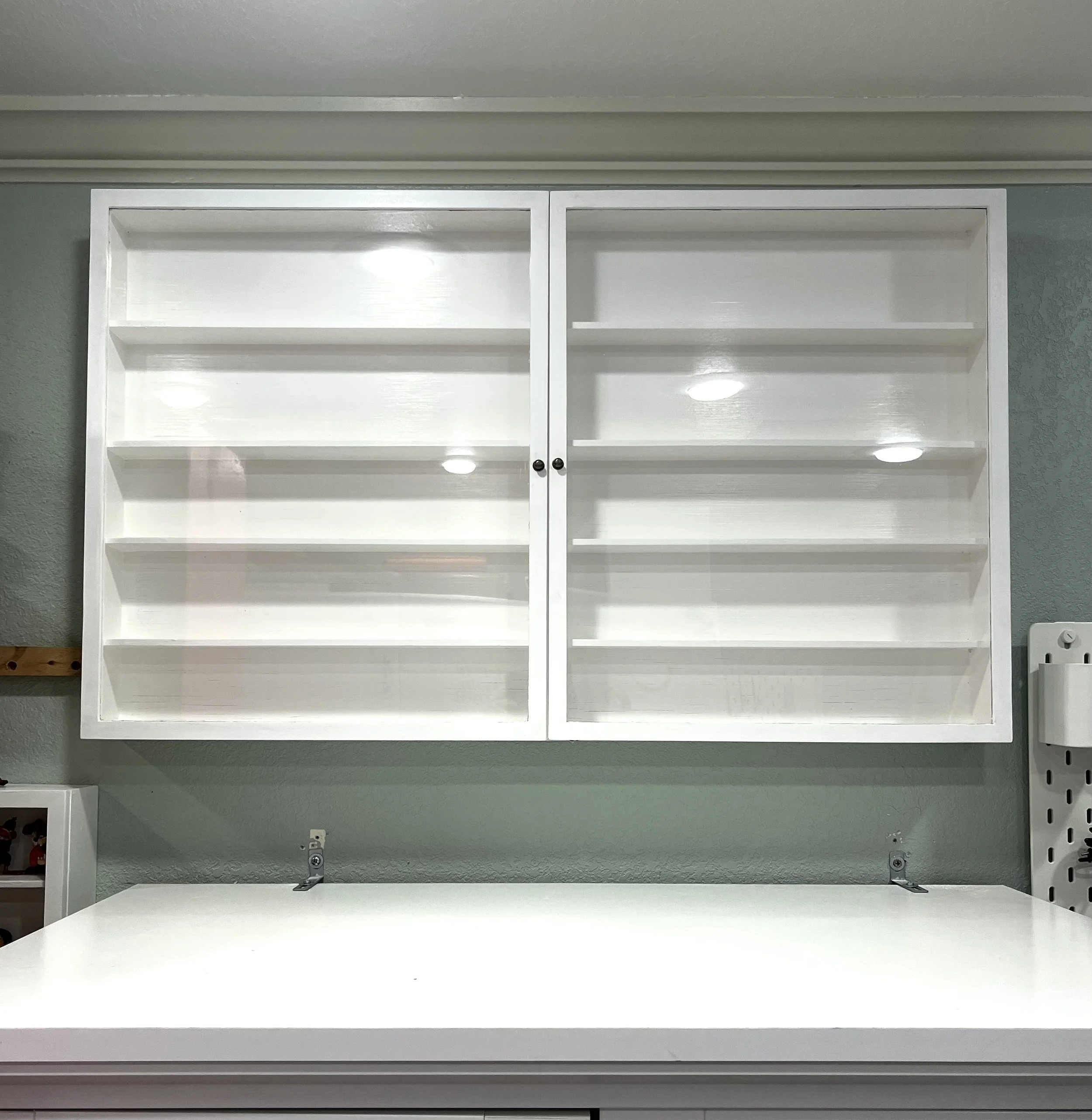 A white bookshelf with two glass doors, empty inside, mounted on a gray wall and illuminated by ceiling lights.