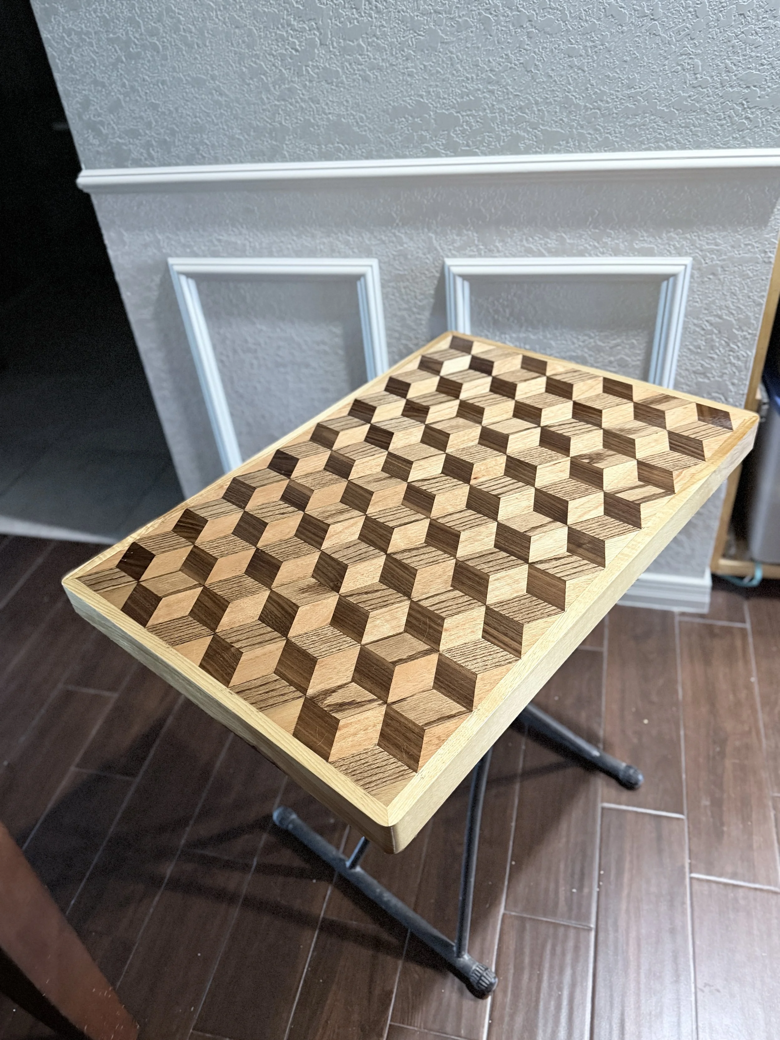 A wooden chessboard with a geometric pattern of light and dark cubes resting on a portable metal table with wheels, in a room with dark brown tiled flooring and a textured light gray wall with decorative molding.