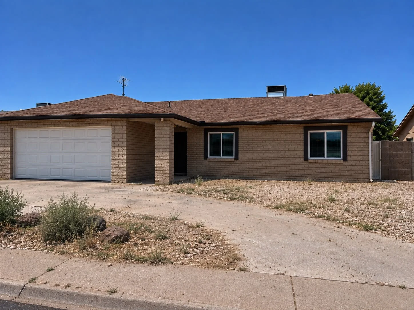 A single-story brick house with a two-car garage, two windows with black shutters, a brown shingled roof, and a front yard with gravel and sparse plants under a clear blue sky.