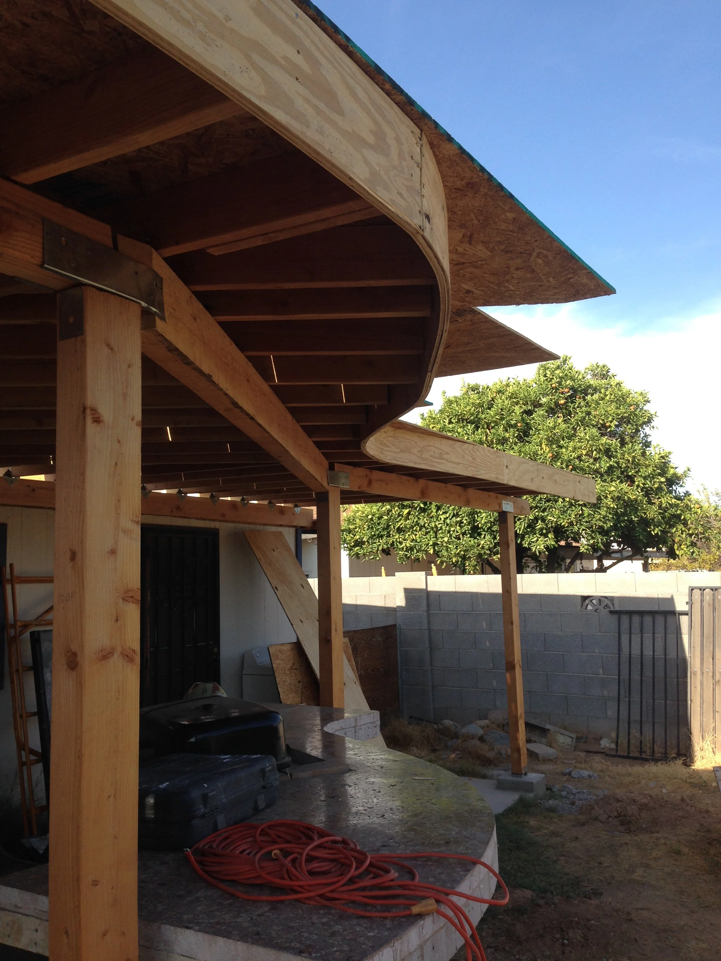 Construction site with wooden framing and a partially built roof, orange extension cord on the ground, and a backyard with trees and a concrete block wall in the background.