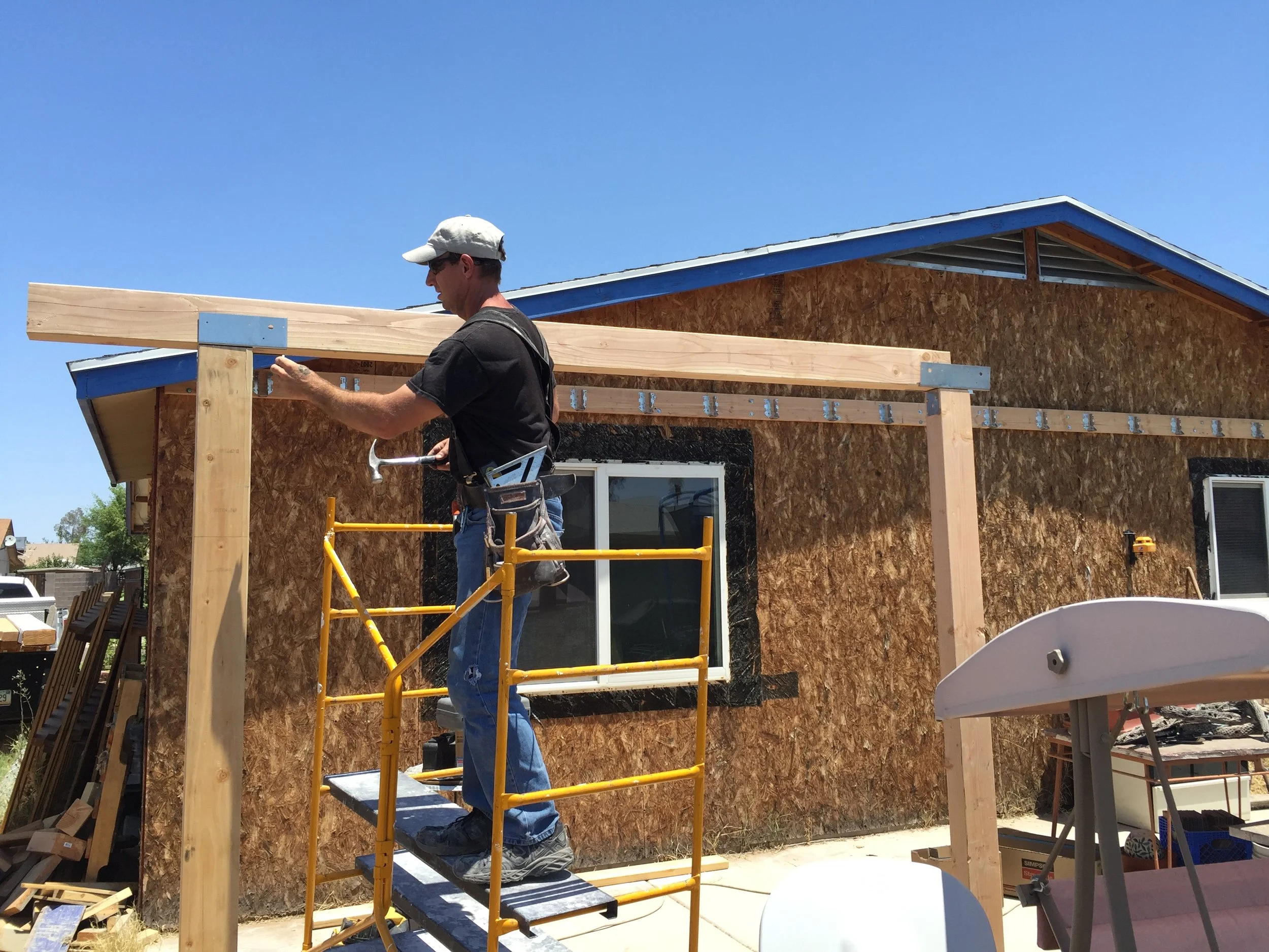 A man wearing a white cap and dark shirt is working on a construction site, standing on a yellow scaffold and hammering a wooden beam attached to a house under construction with a wind hammer. The house has plywood sheathing and a roof with blue trim. The sky is clear and blue.