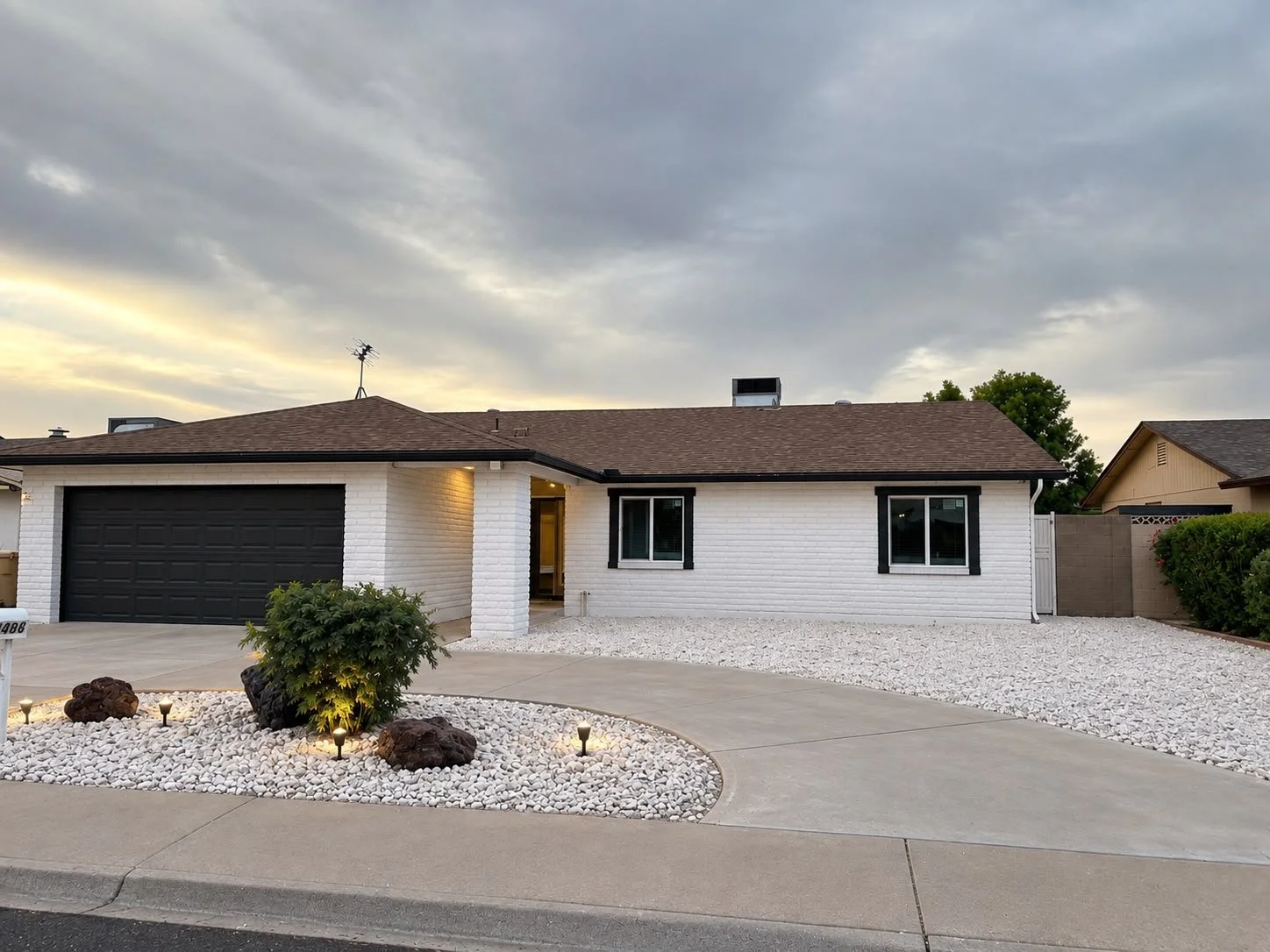 A modern single-story house with a white brick exterior, black window frames, and a black garage door. The front yard features white gravel, a small landscaped area with rocks, a bush, and small lights. The sky is cloudy at dusk.