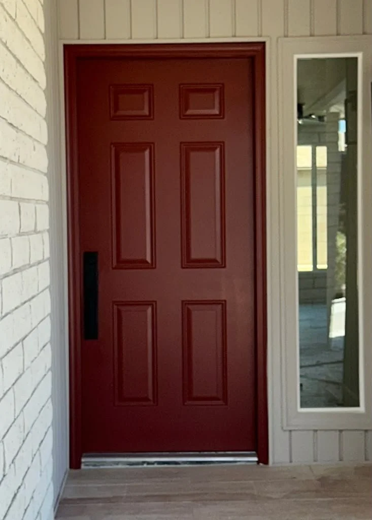 Red front door with four raised rectangular panels, black handle, and a window on the right side showing a porch and screen door outside.