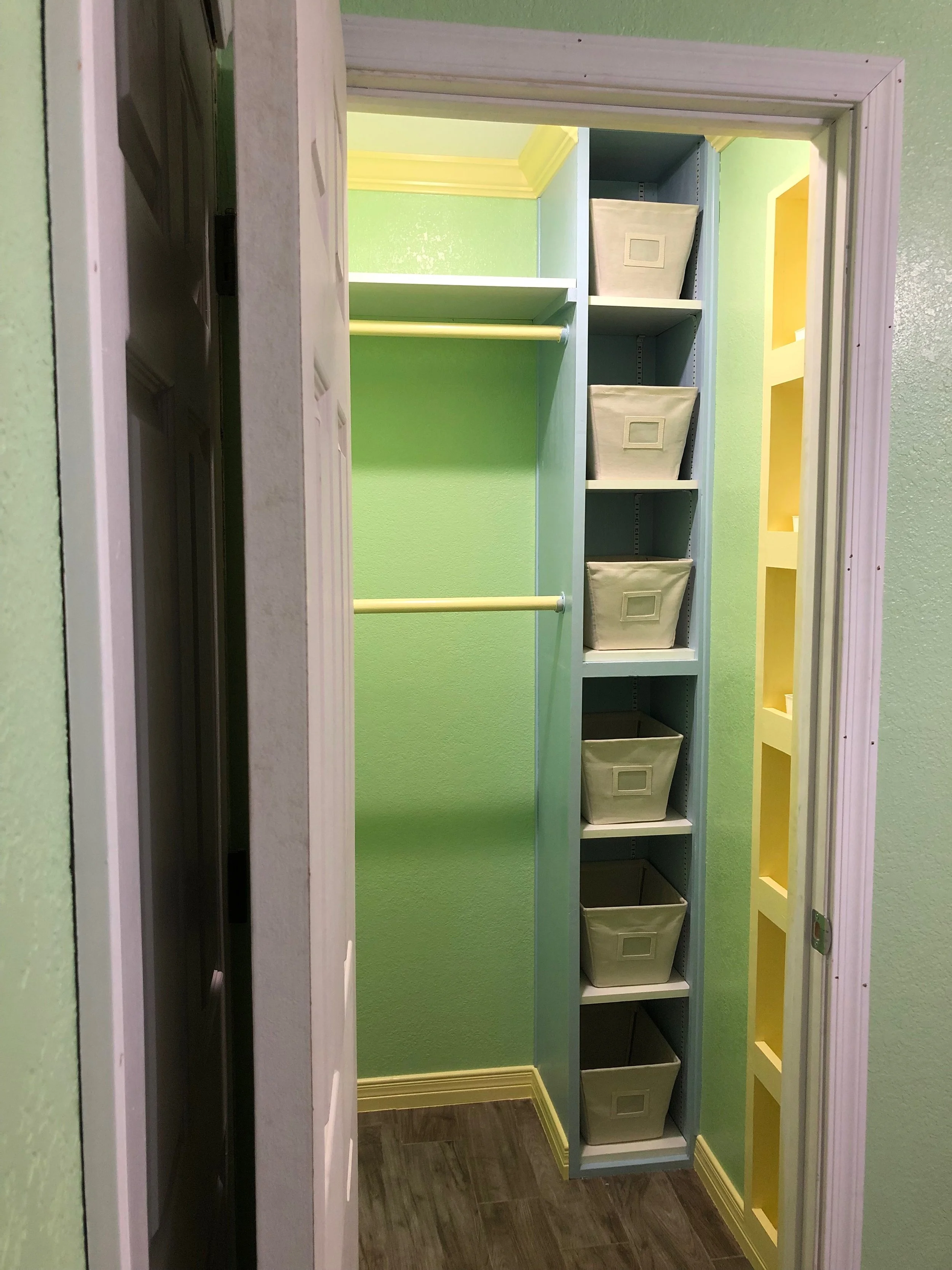 A small closet with green walls and white trim, containing a grey shelving unit with six fabric storage bins, and a yellow built-in cubby on the right wall.
