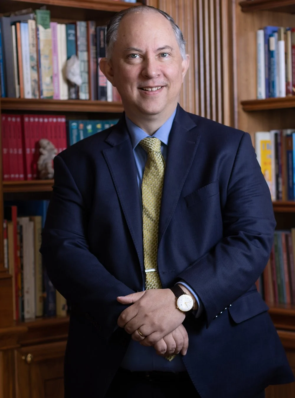 A man in a dark blue suit, light blue shirt, yellow tie, and watch standing in front of a bookshelf filled with colorful books and decorative items.