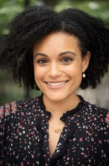 A woman with natural curly hair smiling outdoors, wearing a black floral blouse and pearl earrings.