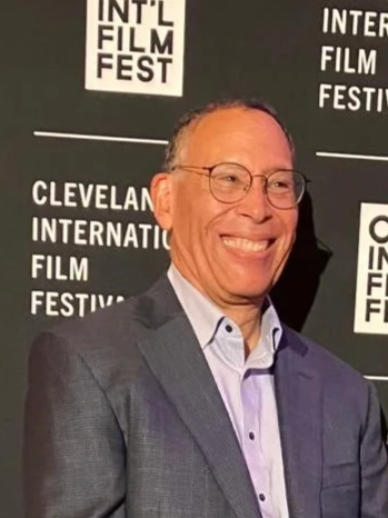 A man in a gray suit and glasses smiling at the Cleveland International Film Festival, standing in front of a black backdrop with festival logos.