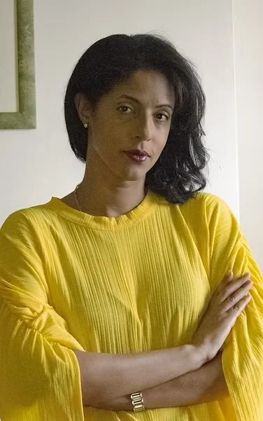 A woman with dark hair wearing a yellow blouse with pleats, standing with her arms crossed in front of a white wall.