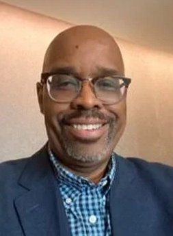 A man wearing glasses, a blue checkered shirt, and a blue blazer, smiling at the camera.
