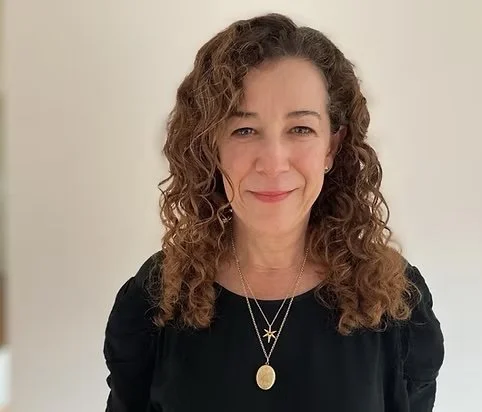 A woman with curly brown hair, smiling, wearing a black top and layered gold necklaces with pendants, standing against a plain neutral background.